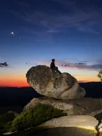 A person sitting on a boulder beneath a crescent moon at sunset, lightly pixelated.
